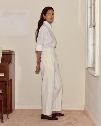 A woman wearing high-waisted Closerie Murray Trousers and a white embroidered shirt stands by a window next to a piano, looking at the camera.