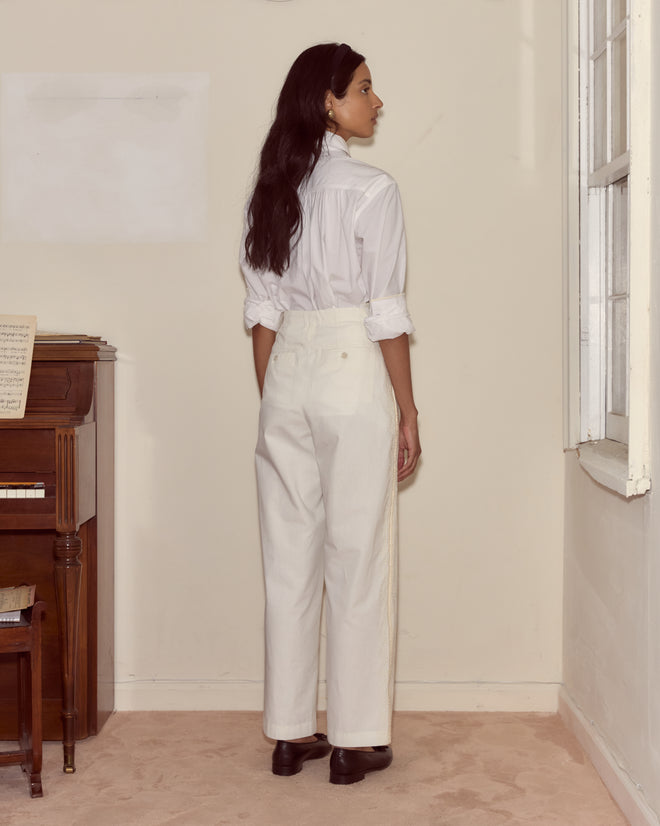 Wearing Closerie Murray Trousers and a white shirt, a woman stands facing a wall near a piano and window in a softly lit room.