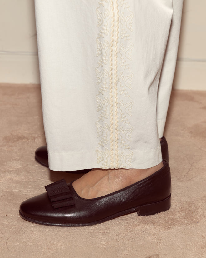 A person wearing Closerie Murray Trousers—high-waisted, wide leg pants with ivory embroidery—and black loafers with a bow, standing on a beige carpet.