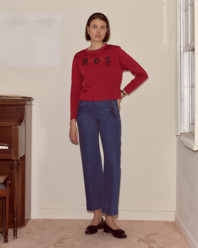 Woman in a Moose Long Sleeve Tee from the Bode collection and blue jeans stands indoors near a piano and window, wearing a neutral expression.