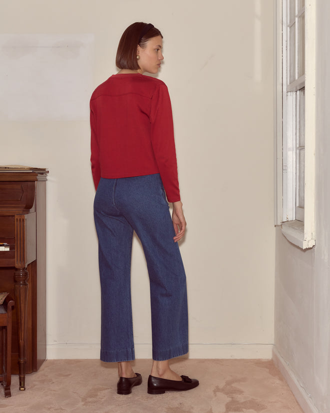 Woman in red Moose Long Sleeve Tee and blue jeans stands indoors, facing away near a window and an upright piano.