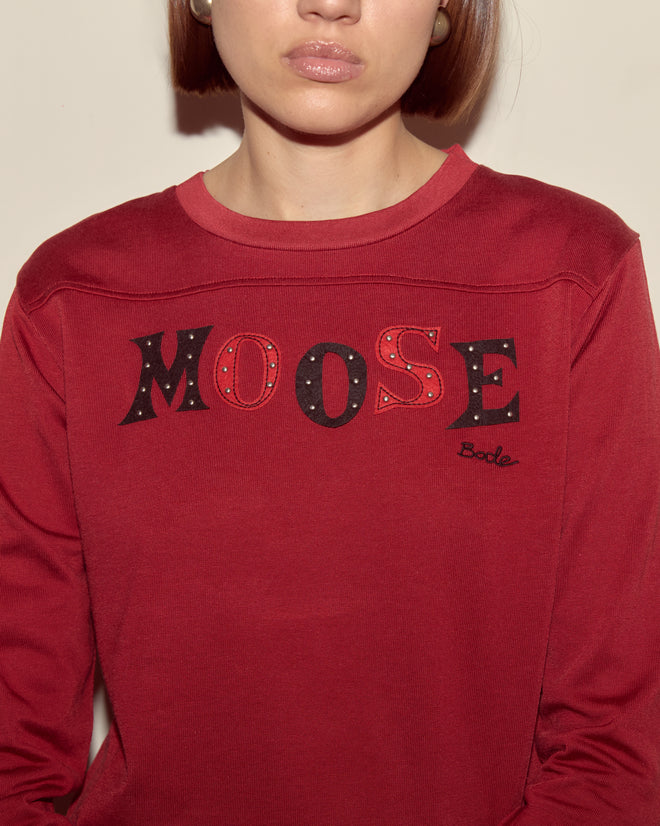 A person wearing the Moose Long Sleeve Tee in red, featuring "MOOSE" and "Bode" embroidery on the front, from the Bode collection.