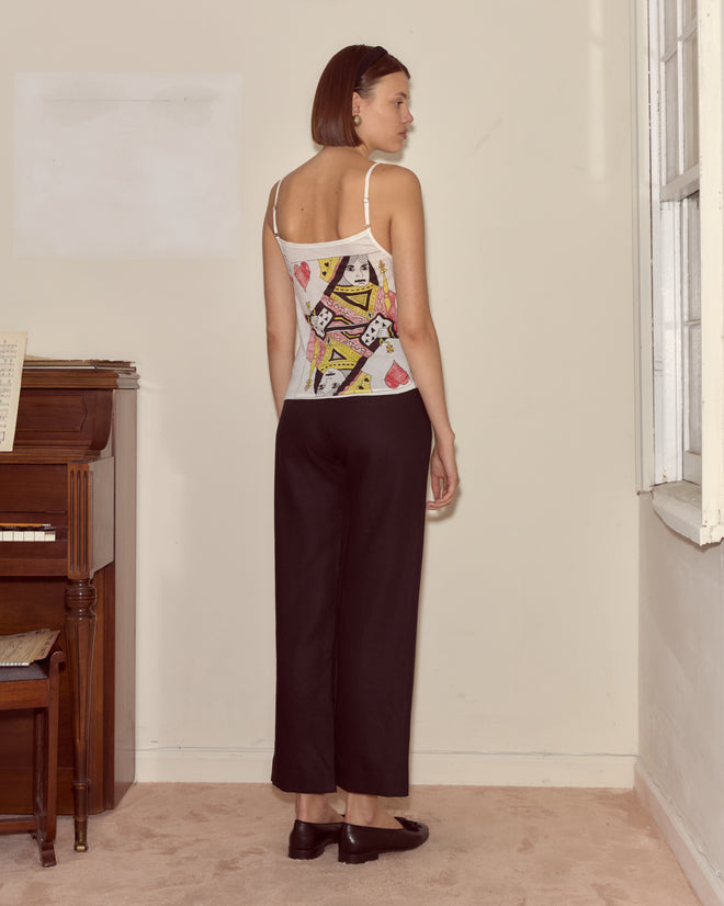 A woman stands indoors facing away in a Queen Of Hearts Tank, black pants, and black shoes near a piano.