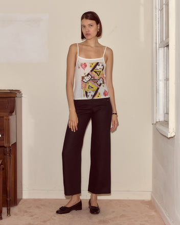 A woman stands indoors by a window, wearing the 100% cotton Queen Of Hearts Tank inspired by Alice Through the Looking Glass, paired with black pants and black shoes.