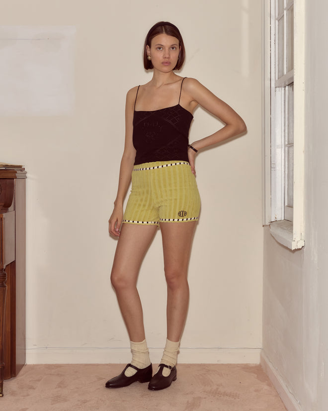 A woman stands indoors by a window wearing the Croquet Ground Shorts—green terrycloth with striped trim—paired with a black camisole, cream socks, and black shoes.