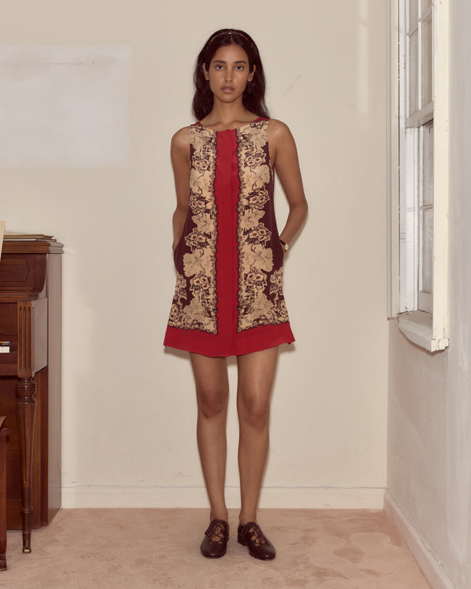 A woman stands indoors wearing the Storybook Dress, a sleeveless, ornate silk mod piece with red accents and pockets, reminiscent of Alice’s Adventures in Wonderland attire.