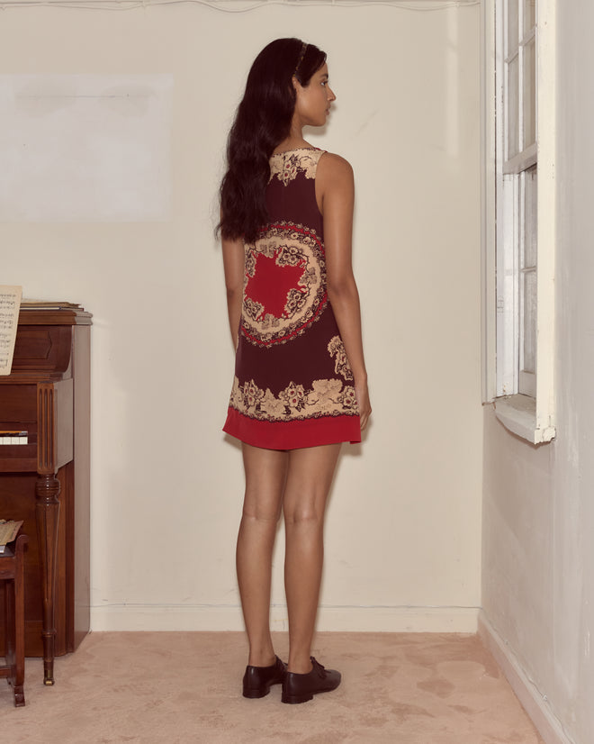 A woman in a Storybook Dress stands facing a wall in a small, sparsely decorated room with a piano.