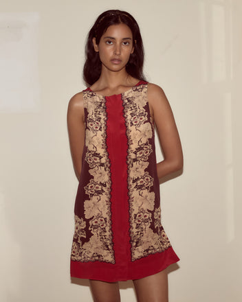 Woman in the Storybook Dress—a sleeveless silk dress featuring ornate beige floral patterns on a red and brown background—standing indoors.