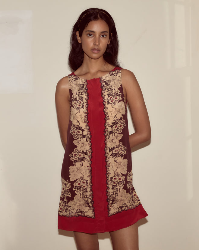 Woman in the Storybook Dress—a sleeveless silk dress featuring ornate beige floral patterns on a red and brown background—standing indoors.