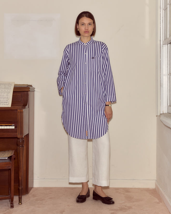 Woman in the Calliope Tunic, a 100% cotton striped top, paired with white pants stands indoors by a piano near a window.