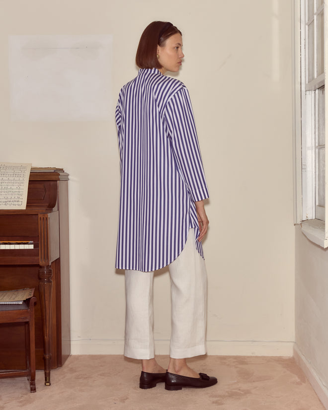 A woman in the Calliope Tunic, featuring 100% cotton and a stripe pattern, pairs it with white pants as she stands by a piano, gazing out of a window.