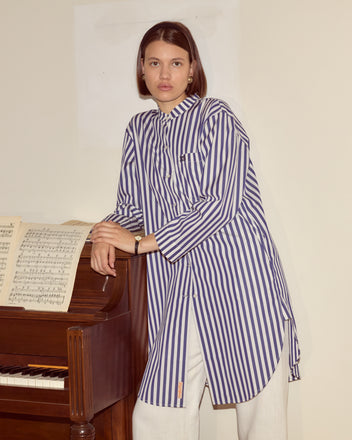 A woman wearing the Calliope Tunic, made of 100% cotton with blue and white stripes, stands beside a piano with sheet music, looking at the camera.
