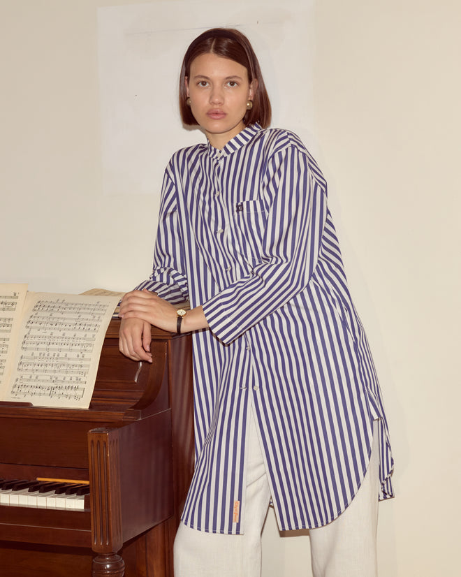A woman wearing the Calliope Tunic, made of 100% cotton with blue and white stripes, stands beside a piano with sheet music, looking at the camera.
