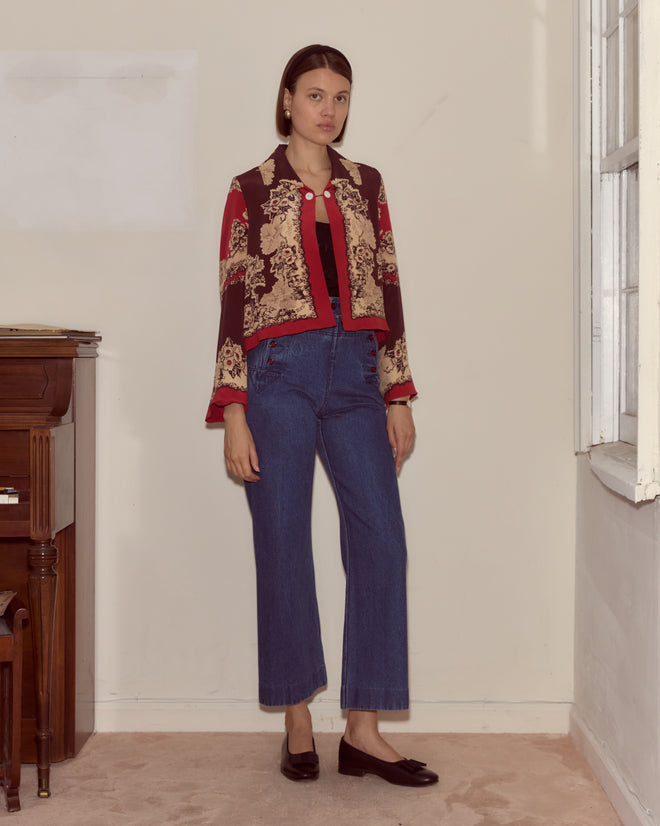 A woman wearing the Storybook Jacket and jeans stands indoors by a window and wooden piano, looking at the camera.