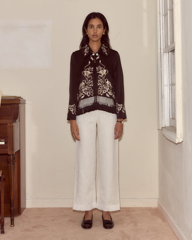 Woman stands indoors wearing the Phantasm Fringe Jacket in black, paired with white wide-leg pants and black shoes.