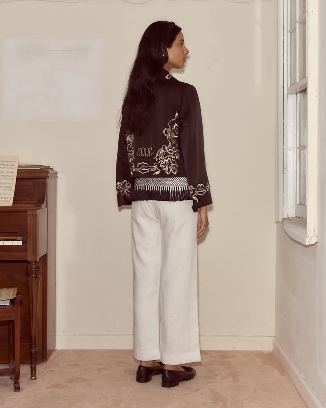 A woman stands indoors near a piano and window, facing away while wearing the Phantasm Fringe Jacket in black paired with white pants.