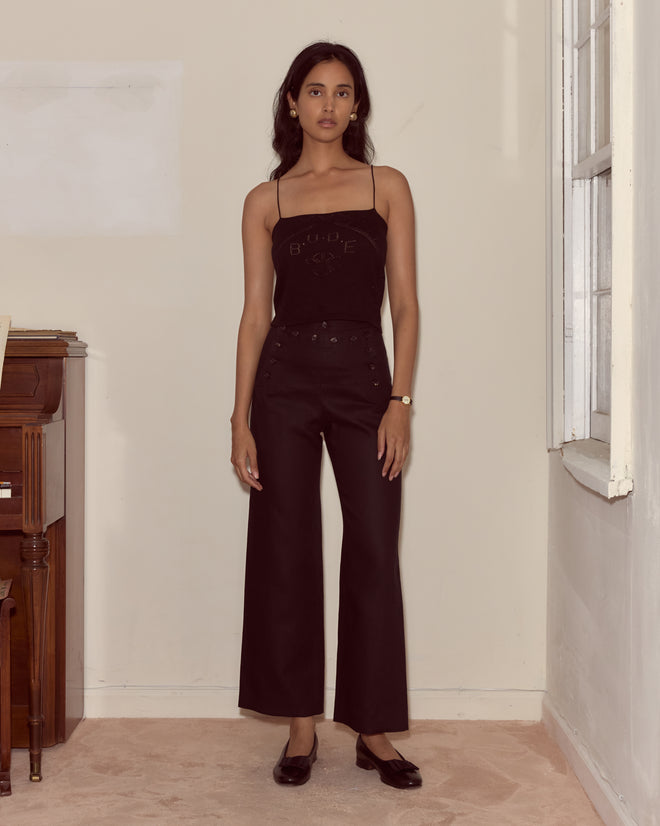 A woman wearing the Anisette Tank and wide-leg pants stands indoors by a piano near a window, facing the camera.