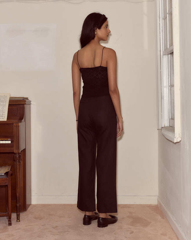 A woman in black pants and the Anisette Tank stands facing a wall in a room with a piano and window.
