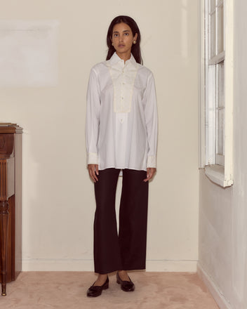 A woman stands indoors against a cream-colored wall, wearing the Closerie Manchester Shirt in white, paired with black pants and black shoes.