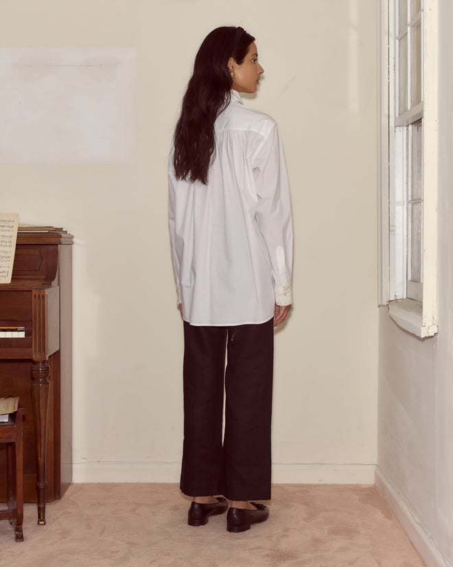 Woman with long dark hair stands indoors, facing a window, wearing the ivory Closerie Manchester Shirt and black pants.