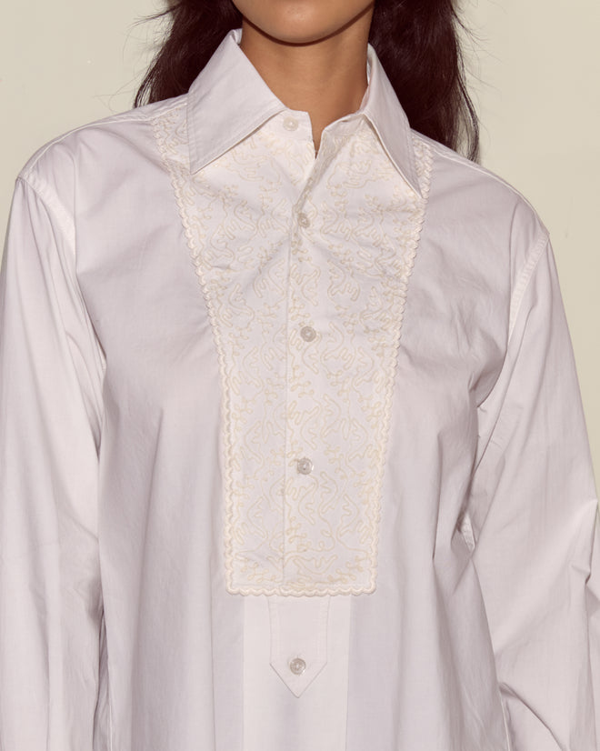 Someone wearing the Closerie Manchester Shirt—an ivory, button-up shirt with decorative embroidery on the front panel.