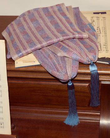 The Cheshire Scarf, featuring handwoven pastel hues with blue tassels and hints of blue and pink plaid, is draped elegantly on a wooden piano with sheet music.