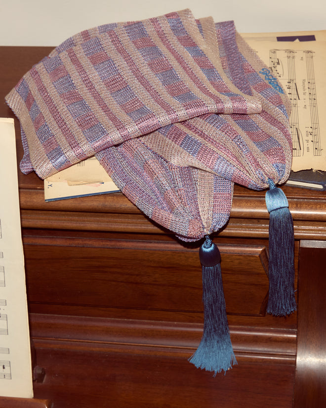 The Cheshire Scarf, featuring handwoven pastel hues with blue tassels and hints of blue and pink plaid, is draped elegantly on a wooden piano with sheet music.