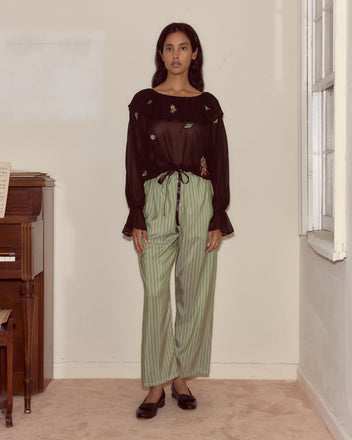A woman in the Flower Toss Blouse and green striped pants stands indoors by a piano and a window.