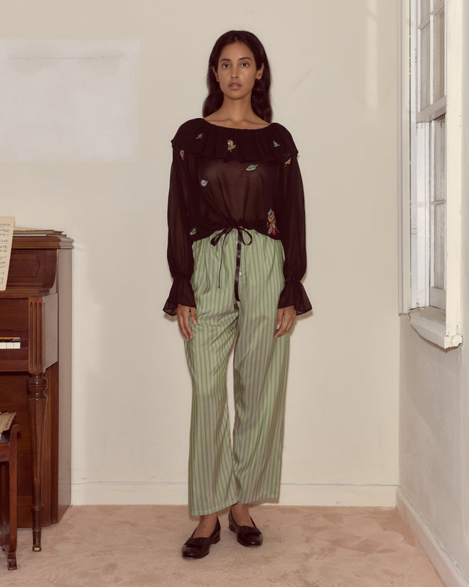A woman in the Flower Toss Blouse and green striped pants stands indoors by a piano and a window.