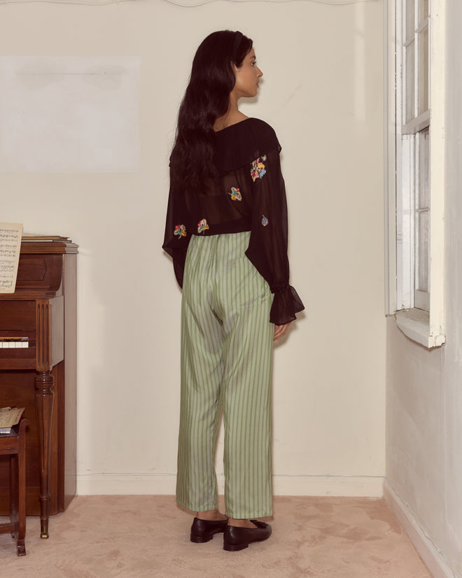 A woman with long dark hair stands indoors facing a wall, wearing green striped pants and the Flower Toss Blouse—a black sheer silk top adorned with delicate floral appliqué.
