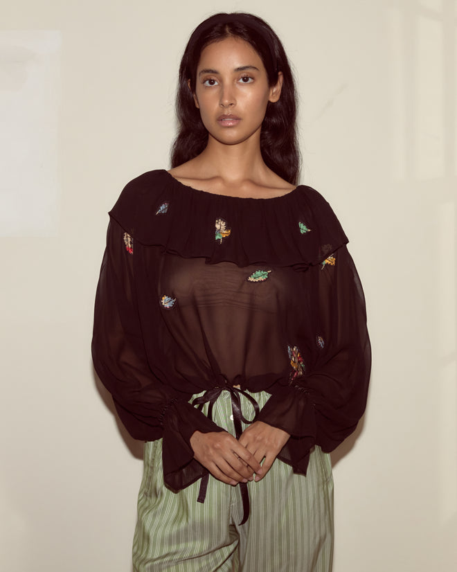 Woman with long dark hair wears the Flower Toss Blouse—a sheer black ruffled top—paired with striped pants, standing against a plain light wall.