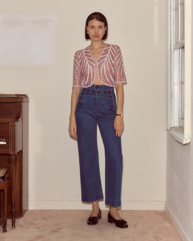 A young woman wears the Beaded Rita Top—a pink and white seed bead design—with high-waisted blue jeans, standing indoors by a piano and window.