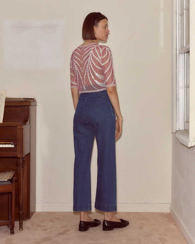 A woman wearing the Beaded Rita Top and blue jeans stands indoors near a window and an upright piano, facing away.