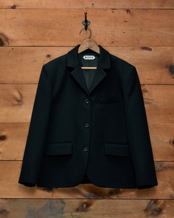 Black Riding Twill Lillian Jacket in heavyweight wool, three-button, boxy fit on a wooden hanger against a wood wall.