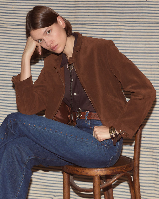 Woman wearing a brown jacket and dark wash jeans sitting on a wooden stool.