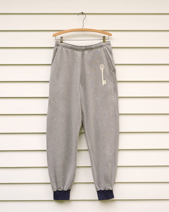 Key Joggers - Grey featuring side pockets and dark cuffs are displayed on a wooden hanger against a white wall—a relaxed style ideal for the Norfolk Chamber Music Festival or a visit to the Yale School of Music.