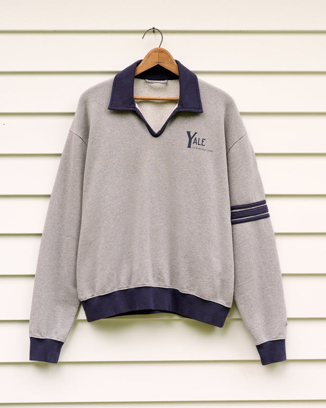 The Yale at Norfolk Polo, a gray sweatshirt featuring a navy collar and cuffs that evoke classic loopback terry polo styling, is displayed on a wooden hanger against a white wall.