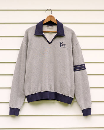 The Yale at Norfolk Polo, a gray sweatshirt featuring a navy collar and cuffs that evoke classic loopback terry polo styling, is displayed on a wooden hanger against a white wall.