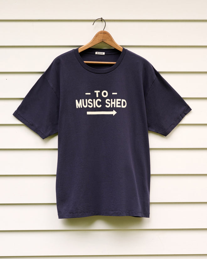Music Shed Tee—navy T-shirt featuring “TO MUSIC SHED” and an arrow, displayed on a hanger against white siding—pays tribute to the iconic Norfolk Music Shed of Yale School of Music’s renowned Norfolk Chamber Music Festival.