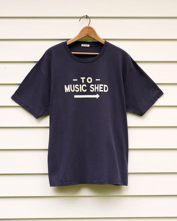Music Shed Tee—navy T-shirt featuring “TO MUSIC SHED” and an arrow, displayed on a hanger against white siding—pays tribute to the iconic Norfolk Music Shed of Yale School of Music’s renowned Norfolk Chamber Music Festival.