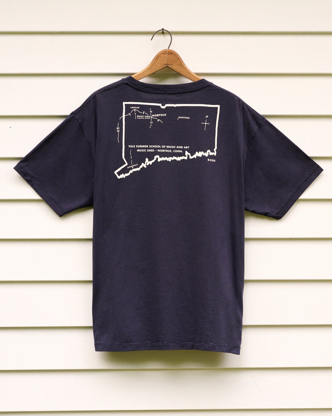 The Music Shed Tee, a navy t-shirt with a white map outline and text referencing the Norfolk Music Shed on the back, is displayed on a hanger against a white paneled wall.