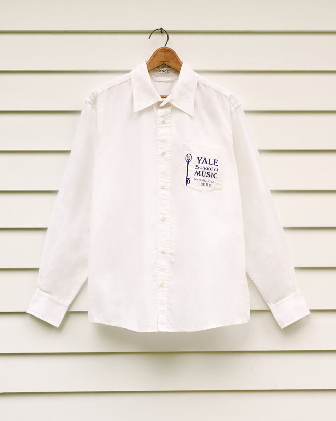 Yale School of Music Shirt in white poplin with a midcentury silhouette, displaying the “Yale School of Music” logo, shown hanging on a wooden hanger against a white wall.