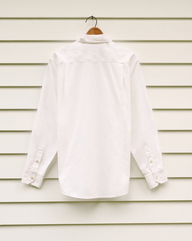 The Yale School of Music Shirt, featuring a plain white poplin design with a midcentury silhouette, hangs on a hanger against a white paneled wall.