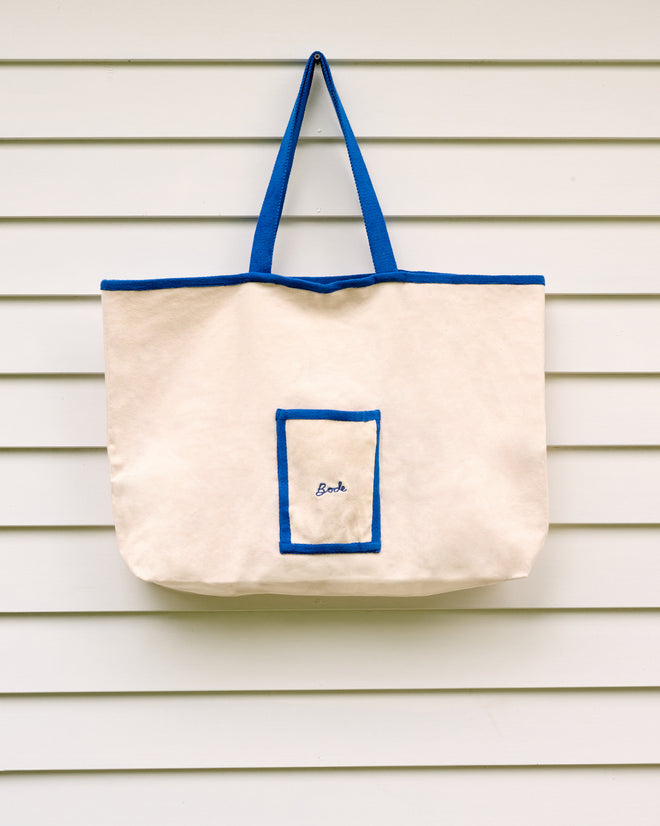 The Yale Postage Tote—a spacious cream bag with blue handles and trim, featuring a “Beach” label—is shown hanging on a white siding wall. This sturdy tote is perfect for carrying your essentials, whether you’re out for the day or attending an event at the Yale School of Music.