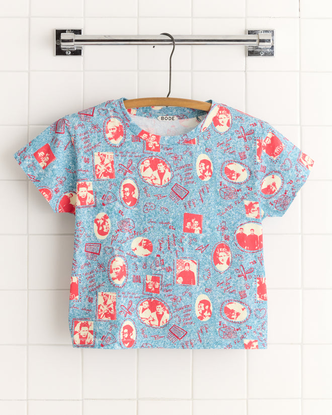 The Yearbook Tee, crafted from 100% cotton, showcases vintage-style red and blue portraits and patterns, presented stylishly on a hanger against a tiled wall.