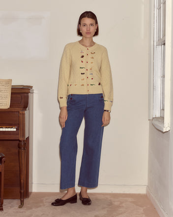 A woman stands indoors by a piano and window, wearing blue jeans and the Zoologist Cardigan—a cream sweater with subtle animal motifs—looking at the camera.