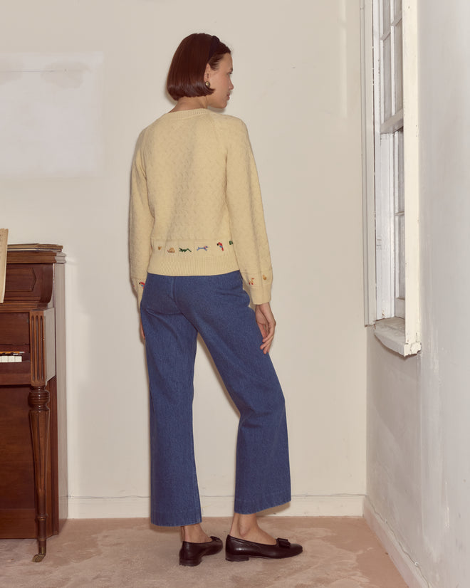 A woman in a Zoologist Cardigan and blue jeans stands facing a wall in a minimalist room featuring a piano and window.