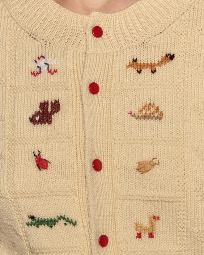 The Zoologist Cardigan is a cream knit sweater featuring red buttons and small animal motifs stitched in colorful yarn on the front.