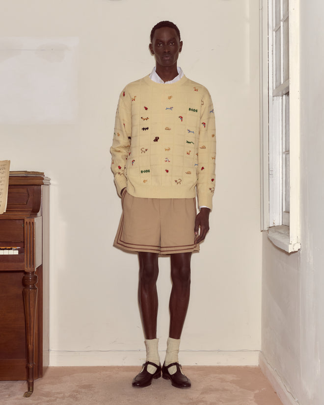 A person in a Zoologist Pullover, tan shorts, socks, and black shoes stands by a piano in a bright room.