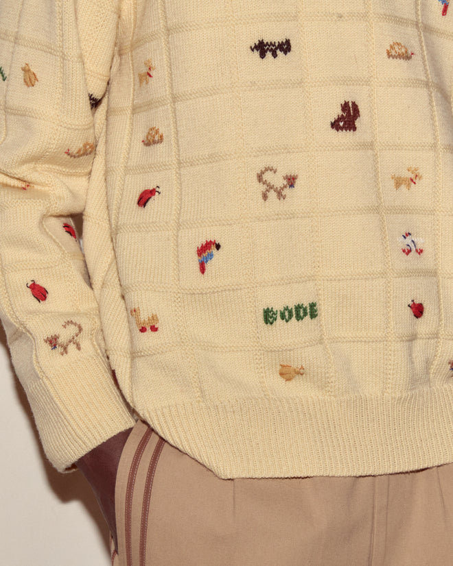 The Zoologist Pullover is a cream knit sweater adorned with small colorful embroidered animals and objects, styled with beige pants featuring brown stripes.